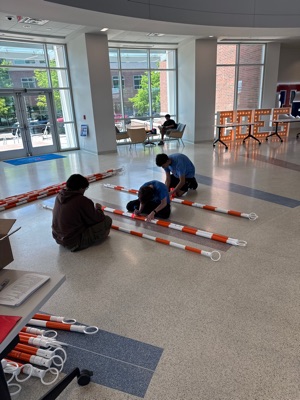 Three students sit on the floor connecting striped poles into rods before assembly begins