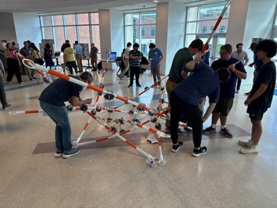 A group of students lifts a partially assembled frame of striped poles off the floor while others hold pieces in place