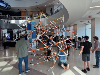 The half-built sculpture rises above onlookers as students work inside its frame, with spectators watching from the second-floor balcony