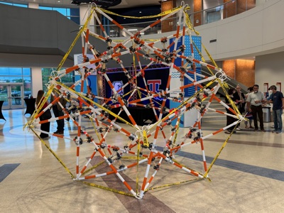 The finished Construction Zone Compound, a large polyhedral sculpture of orange-and-white striped barricade poles and yellow caution tape, standing in the atrium of the engineering building
