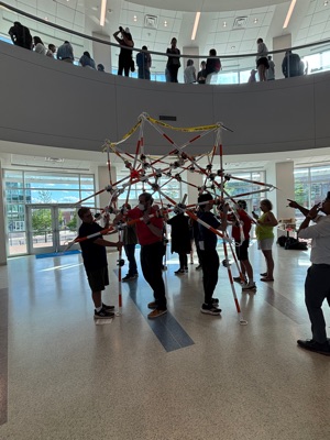 A team of people holds an early tetrahedral frame of poles overhead while assembling it